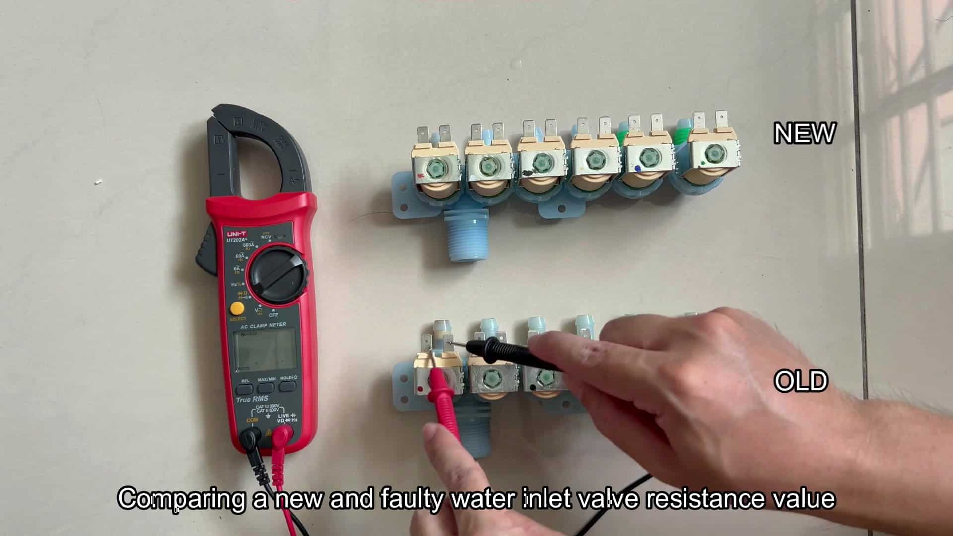A clamp multimeter (brand/model text not clearly readable) shows “OL” on its LCD while red/black probes touch terminals on the lower (labeled “OLD”) multi-solenoid water inlet valve; the upper valve is labeled “NEW,” and a caption reads “Comparing a new and faulty water inlet valve resistance value A clamp multimeter (brand/model text not clearly readable) shows “OL” on its LCD while red/black probes touch terminals on the lower (labeled “OLD”) multi-solenoid water inlet valve; the upper valve is labeled “NEW,” and a caption reads “Comparing a new and faulty water inlet valve resistance value